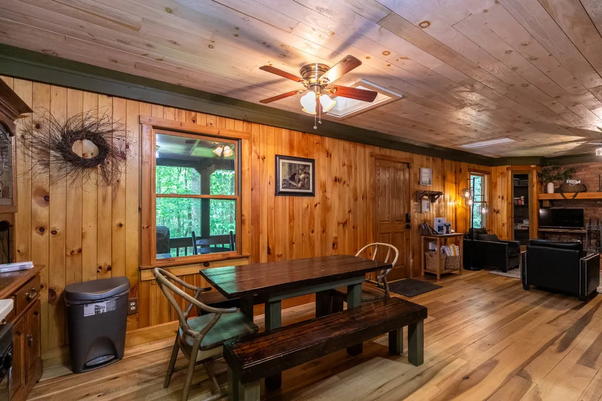 This image showcases a spacious open living area with a large wooden dining table, wood-paneled walls, ceiling fan, prominent windows letting in natural light, and a beautiful fireplace anchoring the space. The room is set up for gatherings, with comfortable seating and a versatile layout that encourages both dining and lounging.