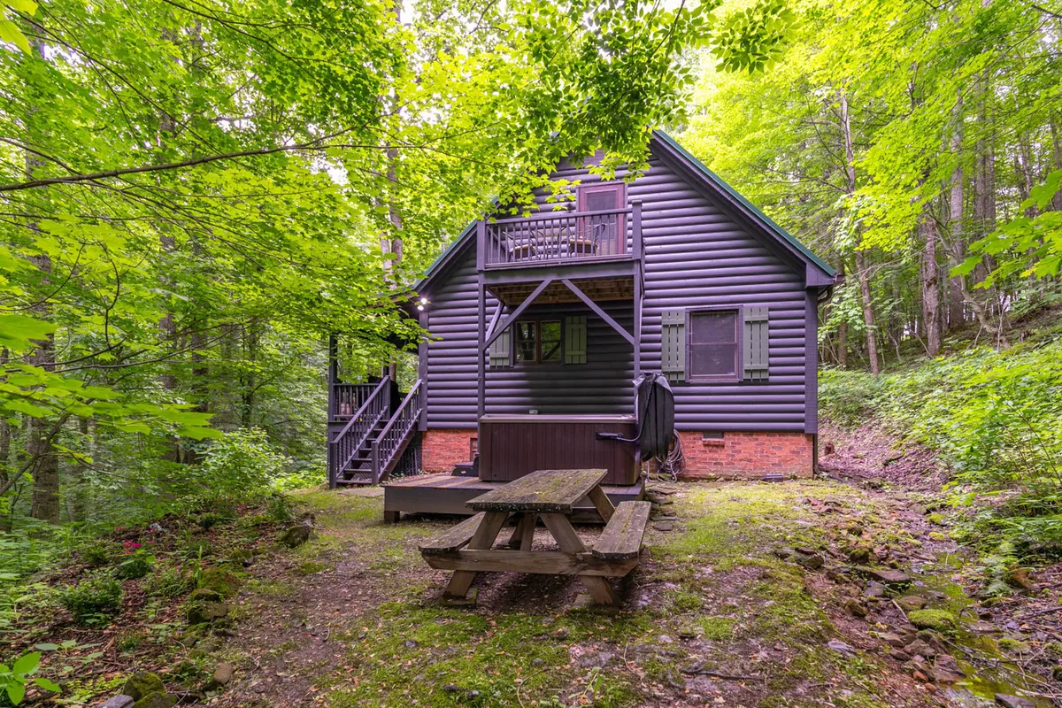 The exterior of a rustic wooden cabin is surrounded by lush greenery, featuring a picnic table perfect for family meals or playing games after a day of exploration. The setting is serene and inviting, emphasizing the cabin’s connection to the outdoors.