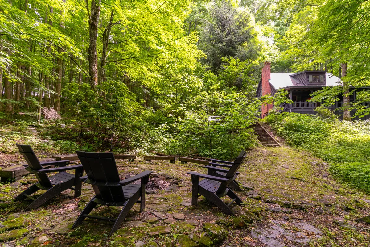 A tranquil outdoor scene features a cozy fire pit surrounded by Adirondack chairs, with a charming cabin nestled among tall trees in the background. The setting is ideal for families to gather, share stories, and enjoy nature together.