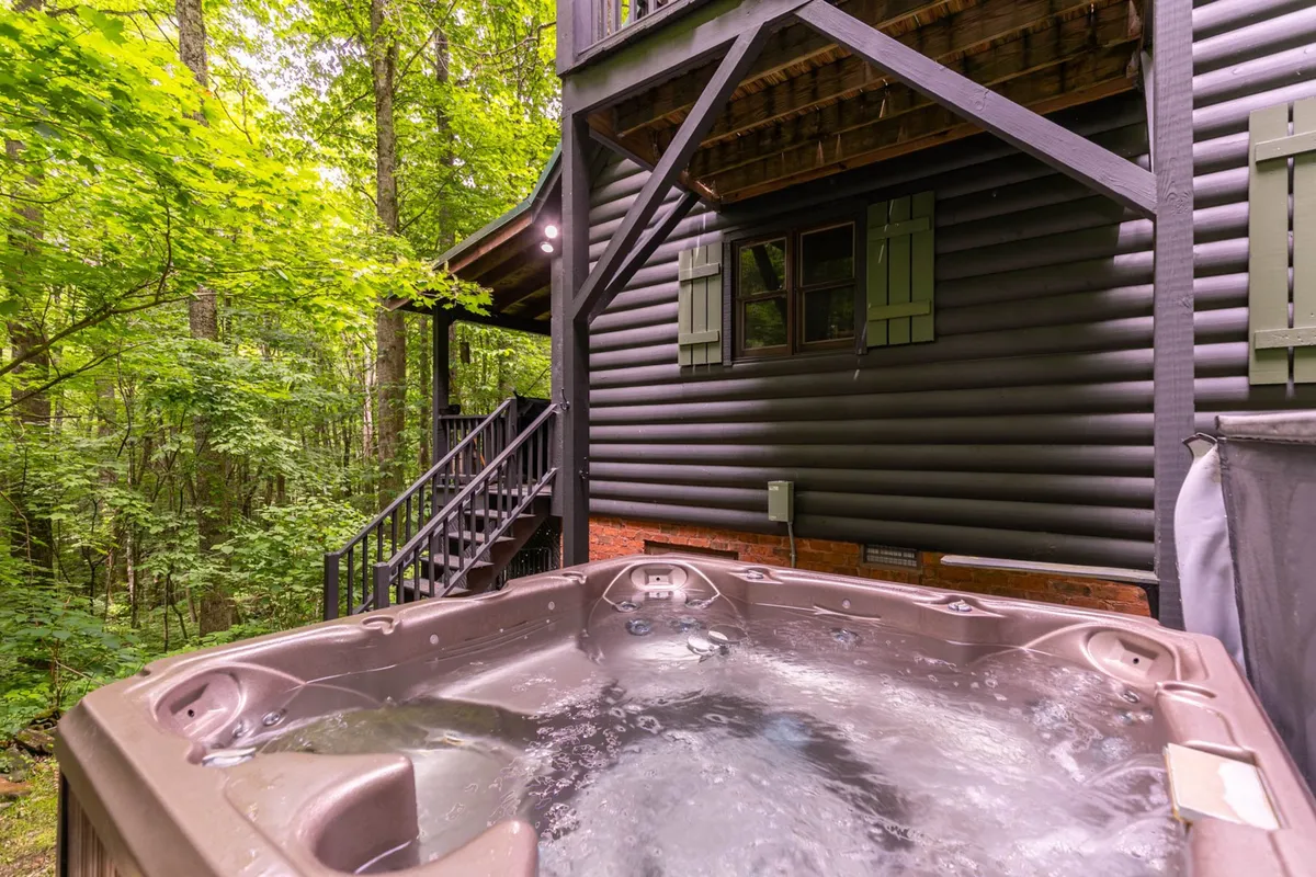 A serene exterior view focused on a hot tub set on a wooden deck, with the cabin’s rustic architecture and stairs leading up to the entrance. The background displays forested tranquility, hinting at evenings spent stargazing or mornings sipping coffee with nature as your backdrop.