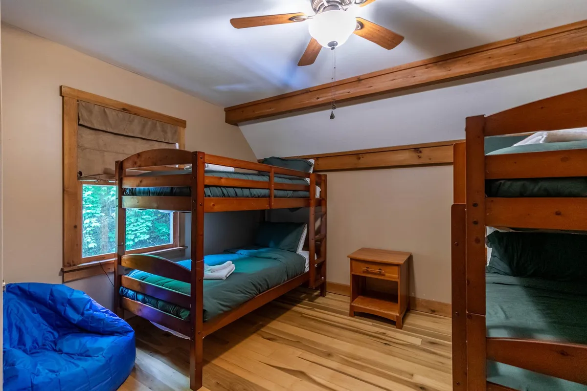 A playful upstairs bunk room featuring two sets of wooden bunk beds, a blue bean bag chair, and a window providing natural light and views of the outdoors. The space is designed for kids or friends, offering both fun and comfort.