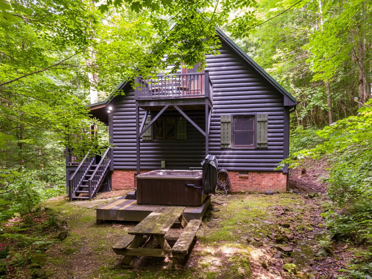 A tranquil wooden deck nestled in lush greenery, featuring a bubbling hot tub, a picnic table, and the log cabin’s rustic exterior. The space is surrounded by forest, evoking privacy and a close-to-nature atmosphere. The scene feels peaceful and inviting, with hints of stargazing, relaxing soaks, and outdoor meals.