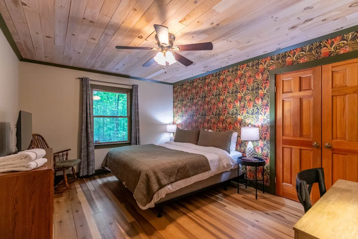 A tranquil bedroom features a bed dressed in neutral bedding, set against a wall adorned with soft floral wallpaper. Natural light pours in from a window, illuminating the peaceful space with its wooden ceiling and flooring. The room evokes a serene, restful mood, ideal for guests seeking relaxation after a busy day.