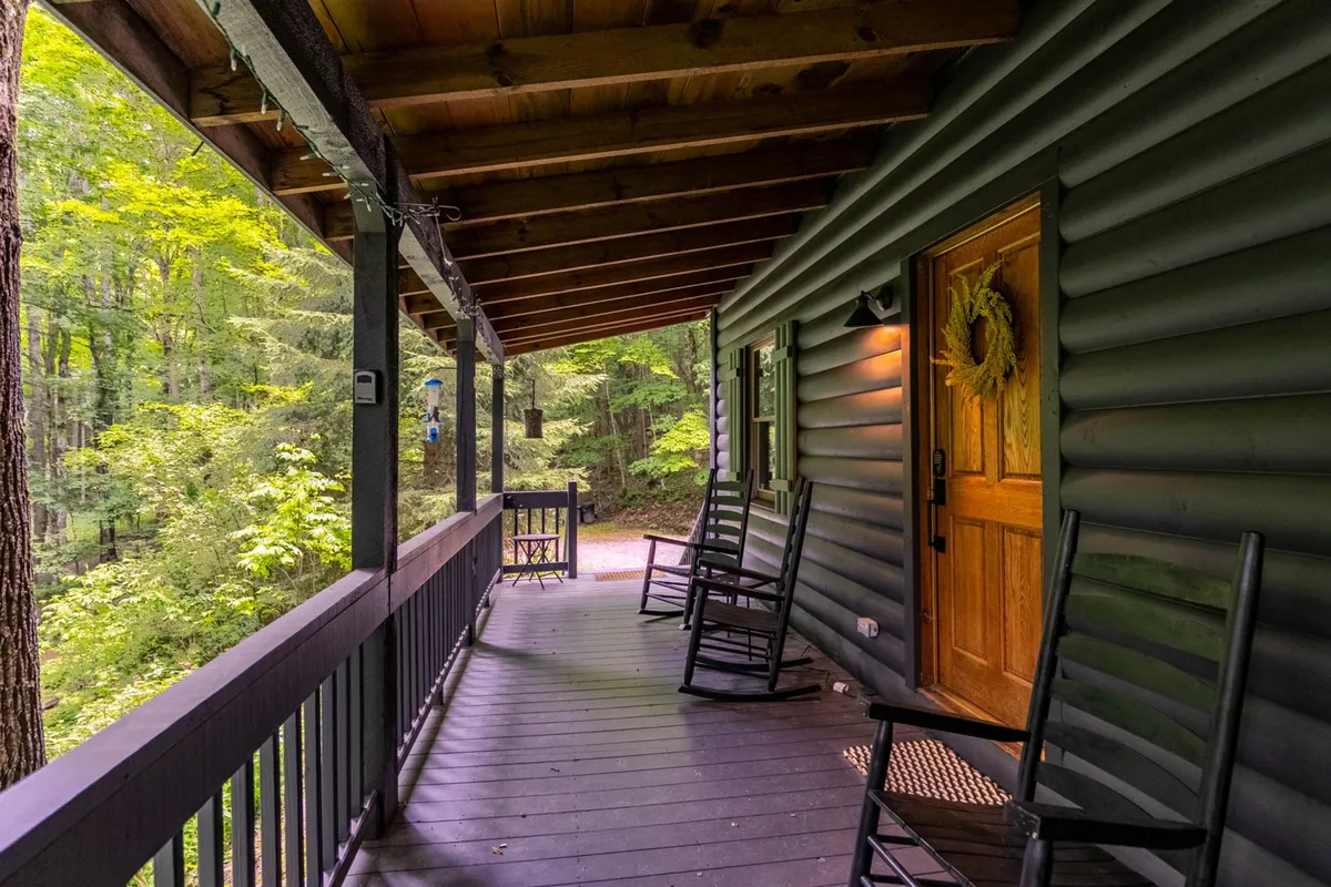 A tranquil porch scene featuring inviting rocking chairs positioned to enjoy the view, a charming wooden front door adorned with a wreath, and lush greenery in the background. The ambiance is one of peace, with the porch offering a perfect spot for wildlife watching or stargazing at night.