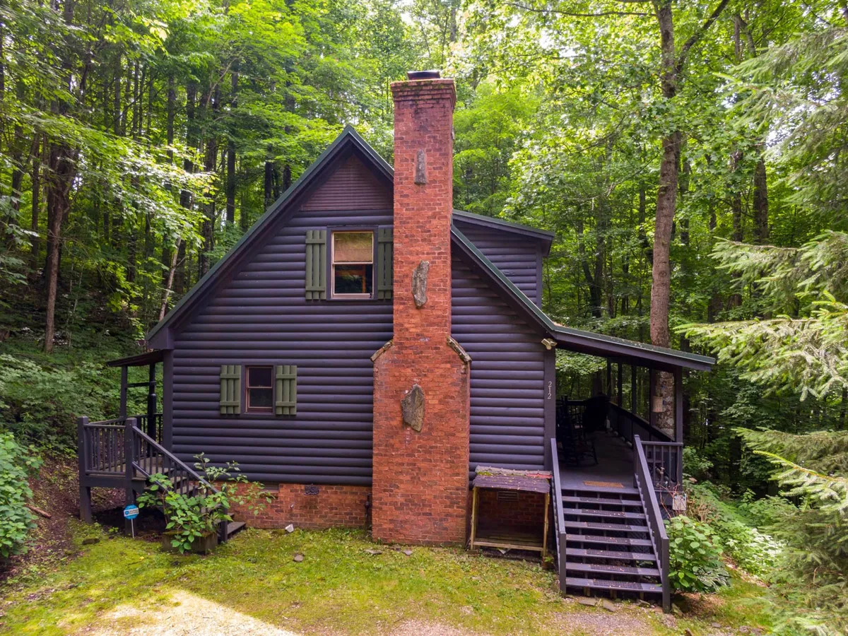 A charming, secluded cabin exterior with a brick chimney, dark wooden siding, and green shutters, surrounded by mature trees. The scene exudes privacy and rustic charm, with the porch offering a quiet spot to enjoy the natural surroundings.