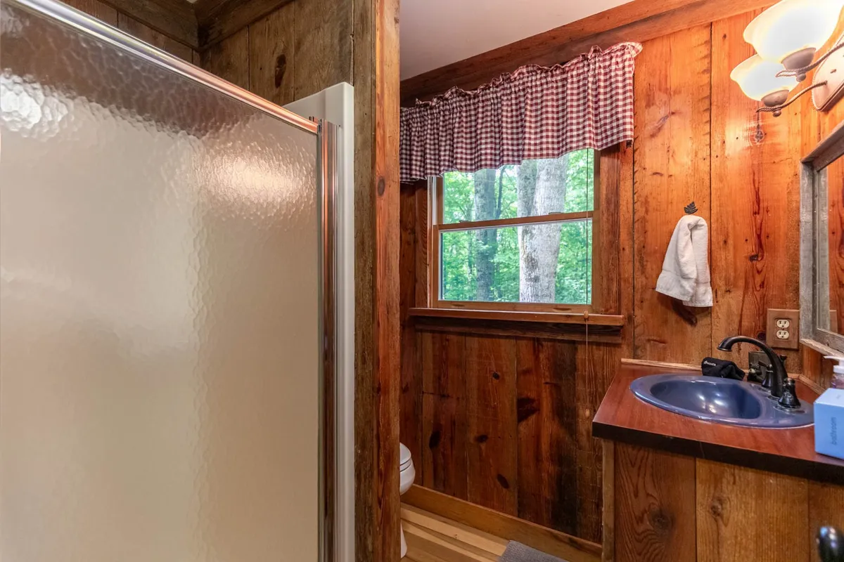 The photo features a warm, wood-paneled bathroom with a blue sink, shower stall, and a window with a checkered curtain. The window provides a peek at the outdoors, while the intimate space suggests a place to refresh after hiking or exploring the area.
